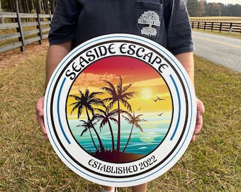May include: A white round sign with a blue and black border. The sign has a sunset scene with palm trees and the text "Seashore Escape Established 2022".