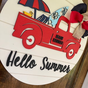 Pu&ograve; includere: Un camioncino rosso di legno con una tavola da surf blu e bianca, un ombrello blu e rosso e un giocattolo giallo nel cassone. Il camioncino ha la scritta "Beach Rentals" sul lato. Il camioncino &egrave; su un cartello rotondo di legno bianco con la scritta "Hello Summer" in nero.