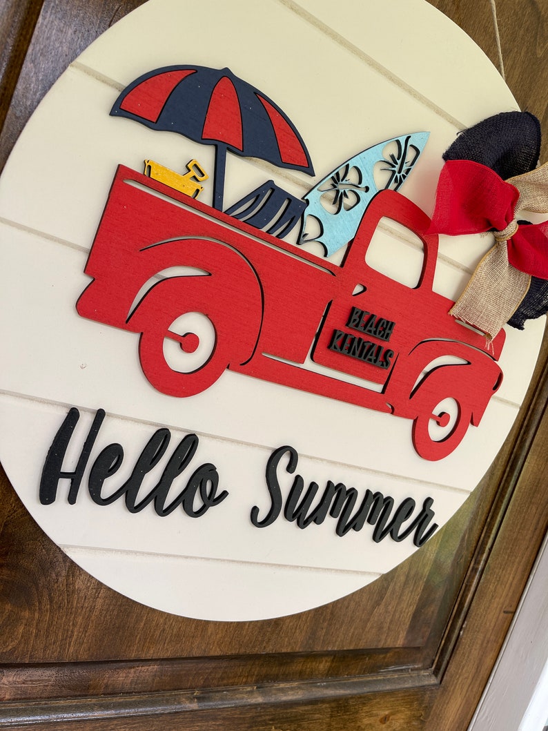 Pu&ograve; includere: Un camioncino rosso di legno con una tavola da surf blu e bianca, un ombrello blu e rosso e un giocattolo giallo nel cassone. Il camioncino ha la scritta "Beach Rentals" sul lato. Il camioncino &egrave; su un cartello rotondo di legno bianco con la scritta "Hello Summer" in nero.