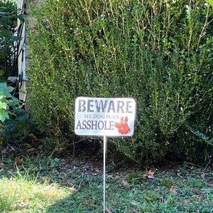 K&ouml;nnte beinhalten: Ein wei&szlig;es Schild mit schwarzem Text, auf dem "BEWARE MY DOG IS AN ASSHOLE" steht, mit einer Cartoon-Hundeillustration. Das Schild ist auf einem Metallpfahl und steht vor einem gr&uuml;nen Busch.