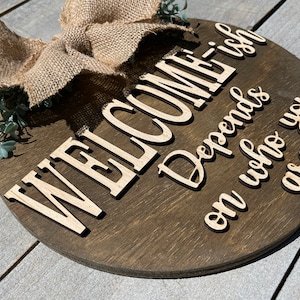 May include: A round wooden sign with the text "WELCOME Depends on who you are -ish" in white lettering. The sign is decorated with a burlap bow and green foliage.