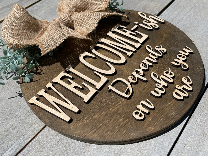 May include: A round wooden sign with the text "WELCOME Depends on who you are -ish" in white lettering. The sign is decorated with a burlap bow and green foliage.