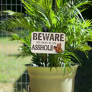 K&ouml;nnte beinhalten: Ein gr&uuml;ner Pflanzentopf mit einem Schild, auf dem "BEWARE MY DOG IS AN ASSHOLE" steht, mit einem Cartoon-Hund darauf.