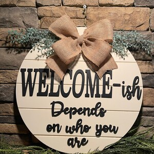 May include: A round wooden sign with the text "WELCOME-ish Depends on who you are" in black lettering. The sign is decorated with a burlap bow and greenery.