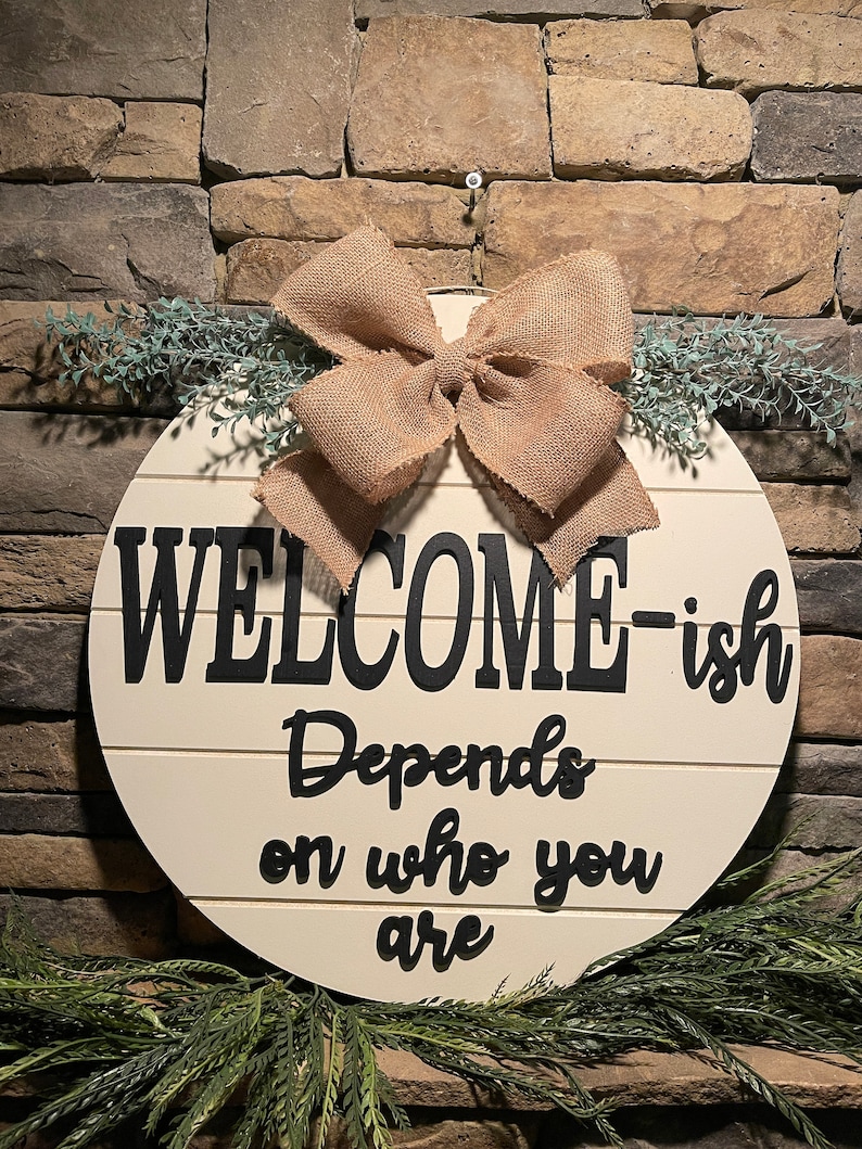 May include: A round wooden sign with the text "WELCOME-ish Depends on who you are" in black lettering. The sign is decorated with a burlap bow and greenery.
