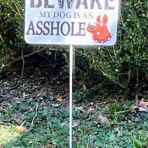K&ouml;nnte beinhalten: Ein wei&szlig;es Schild mit schwarzem Text, auf dem "BEWARE MY DOG IS AN ASSHOLE" steht. Ein roter Cartoon-Hund mit einem wei&szlig;en Halsband befindet sich in der unteren rechten Ecke des Schildes. Das Schild ist an einem Metallpfahl befestigt und befindet sich in einem grasbewachsenen Bereich.
