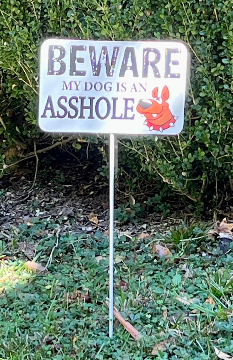 K&ouml;nnte beinhalten: Ein wei&szlig;es Schild mit schwarzem Text, auf dem "BEWARE MY DOG IS AN ASSHOLE" steht. Ein roter Cartoon-Hund mit einem wei&szlig;en Halsband befindet sich in der unteren rechten Ecke des Schildes. Das Schild ist an einem Metallpfahl befestigt und befindet sich in einem grasbewachsenen Bereich.