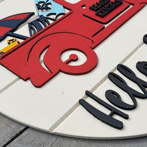 Pu&ograve; includere: Un cartello rotondo in legno bianco con un camioncino rosso che trasporta oggetti da spiaggia e il testo "Hello Summer" in lettere nere.