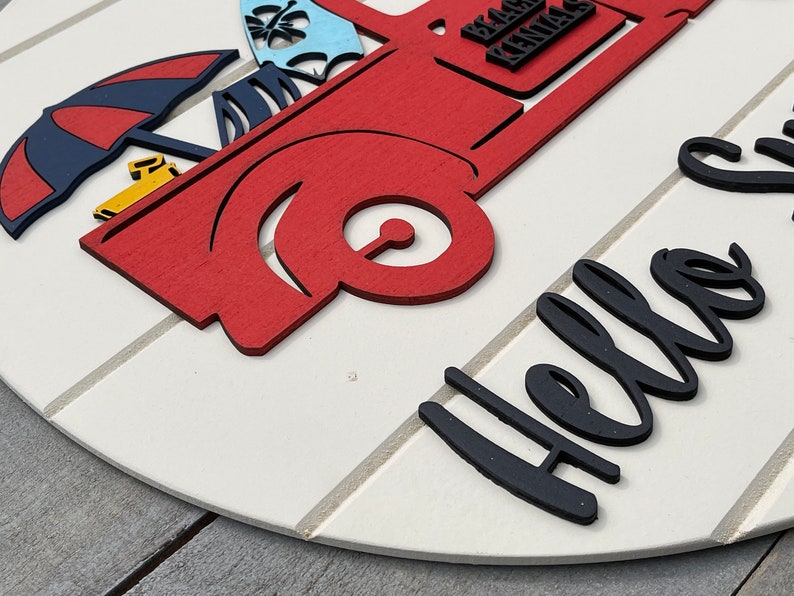 Pu&ograve; includere: Un cartello rotondo in legno bianco con un camioncino rosso che trasporta oggetti da spiaggia e il testo "Hello Summer" in lettere nere.