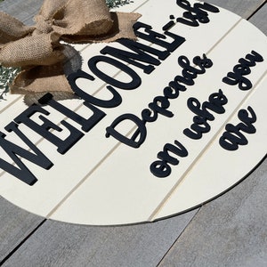 May include: White round wooden sign with black lettering that reads "Welcome is Depends on who you are". The sign has a burlap bow and greenery on top.