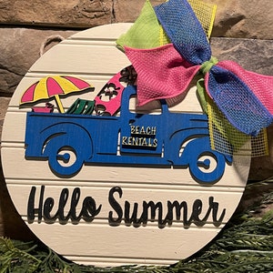 Pu&ograve; includere: Un cartello rotondo in legno bianco con un camioncino blu che trasporta un ombrellone da spiaggia, sedie da spiaggia e tavole da surf. Il camioncino ha la scritta "Beach Rentals" sul lato. Il cartello ha anche la scritta "Hello Summer" in lettere nere e un fiocco rosa, verde e blu.
