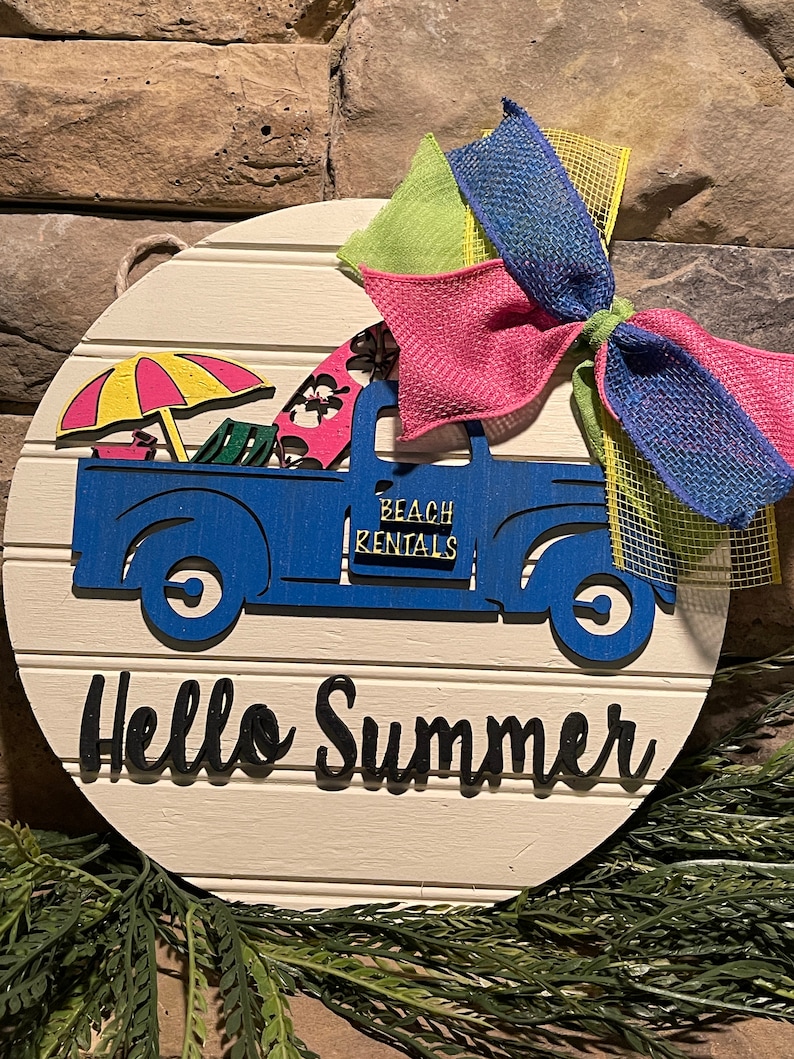 Pu&ograve; includere: Un cartello rotondo in legno bianco con un camioncino blu che trasporta un ombrellone da spiaggia, sedie da spiaggia e tavole da surf. Il camioncino ha la scritta "Beach Rentals" sul lato. Il cartello ha anche la scritta "Hello Summer" in lettere nere e un fiocco rosa, verde e blu.