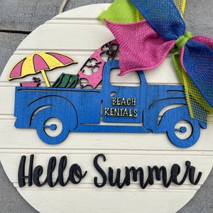 Pu&ograve; includere: Un cartello rotondo in legno con un camioncino blu che trasporta tavole da surf, un ombrello e sedie da spiaggia. Il camioncino ha la scritta "Beach Rentals" sul lato. Il cartello ha anche le parole "Hello Summer" in lettere nere.