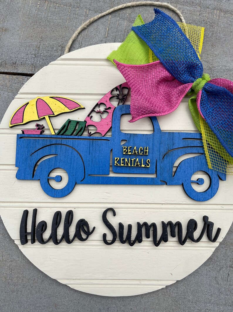 Pu&ograve; includere: Un cartello rotondo in legno con un camioncino blu che trasporta tavole da surf, un ombrello e sedie da spiaggia. Il camioncino ha la scritta "Beach Rentals" sul lato. Il cartello ha anche le parole "Hello Summer" in lettere nere.