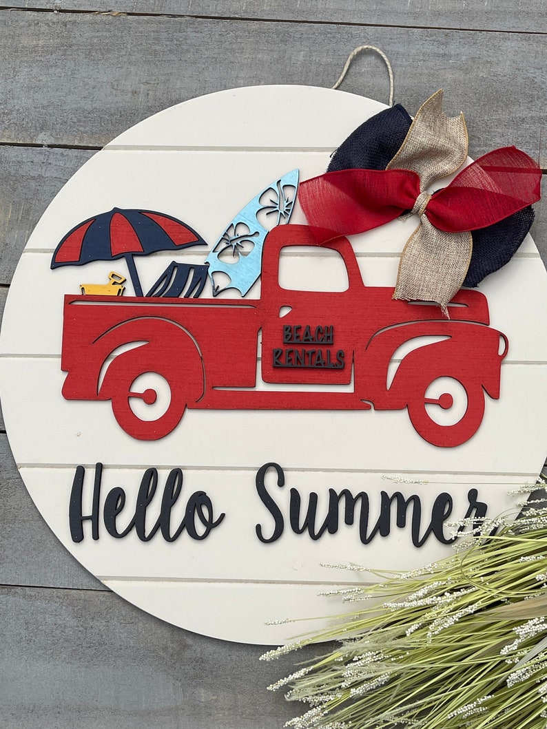 Pu&ograve; includere: Camioncino rosso in legno con una tavola da surf, un ombrellone da spiaggia e sedie da spiaggia nel cassone. Il camioncino ha la scritta "Beach Rentals" sul lato. Il camioncino &egrave; su un cartello rotondo in legno bianco con la scritta "Hello Summer" scritta sotto. Un fiocco rosso, bianco e marrone &egrave; attaccato alla parte superiore del cartello.