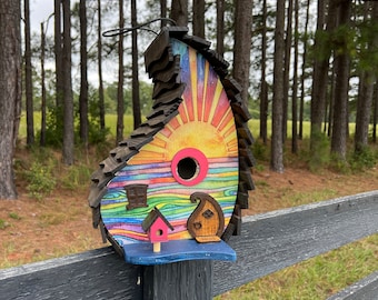May include: A colorful wooden birdhouse shaped like a teardrop with a rainbow design, a sun, and a small pink house. The birdhouse is mounted on a black wooden fence post.