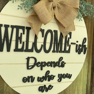 May include: A white round wooden sign with the text "Welcome-ish Depends on who you are" in black lettering. The sign is decorated with a burlap bow and green sprigs.