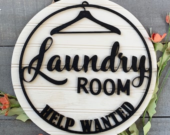 May include: A white round wooden sign with black lettering that reads "Laundry Room Help Wanted". The sign has a black hanger silhouette at the top.