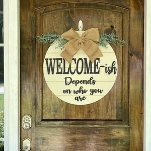 May include: A round wooden sign with the text "Welcome-ish Depends on who you are" in black lettering. The sign is decorated with a burlap bow and greenery.