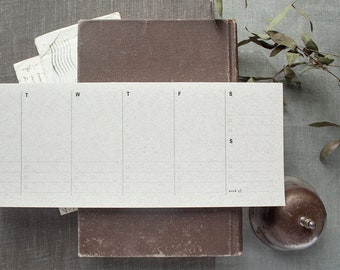 May include: A weekly planner with a simple design. The planner has seven columns for each day of the week, Monday through Sunday. The days of the week are labelled at the top of each column. The planner is printed on white paper with a subtle texture.