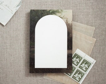 May include: A white card with an arched top, sitting on a postcard with a green and brown landscape. The card is blank and ready for writing.