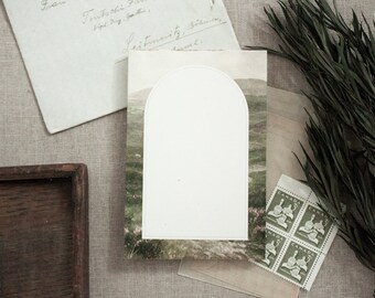 May include: A blank postcard with a white archway shape in the centre. The postcard has a green and brown landscape image with a blurry background.