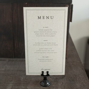May include: A brown paper menu card with black text. The menu card is titled 'MENU' and lists the following items: 'to start', 'Italian antipasto bar', 'spinach salad with julienne of pear, asiago cheese, dried cranberries tossed in zesty lemon pepper', 'entrée', 'boneless skin-on chicken breast served with rice pilaf and creole butter', 'grilled salmon with pickled mustard salsa verde, seasonal vegetables and potato pavé', 'late night', 'homemade Italian pizza and poutine station', 'bon appétit'. The menu card is displayed on a black metal stand.
