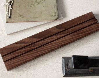 May include: A dark brown wooden desk organiser with three grooves. The organiser is rectangular and has a natural wood grain. A vintage notebook with the word "Autographs" is in the background, along with a small notepad and a metal object.