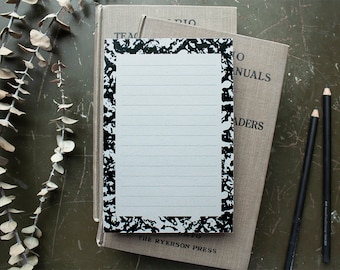 May include: A lined notepad with a black and white speckled border rests on two vintage books. Two black pencils are positioned to the right. Dried eucalyptus branches are draped on the left, creating a rustic, artistic composition.