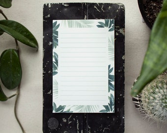 May include: A white notepad with a green and white tropical leaf border. The notepad is on a black surface.