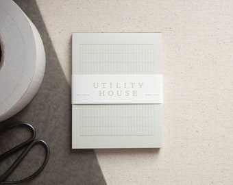May include: A white notepad with a grid pattern and the text "UTILITY HOUSE EST 2018 RULE-DOT" printed on the front.