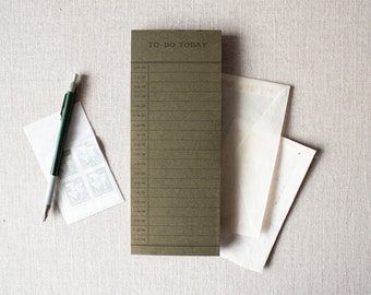 May include: An olive green To-Do list notepad with time slots, a green and silver pen, stamps, and envelopes. The notepad has the text "TO-DO TODAY" at the top. The items are arranged on a neutral linen surface.