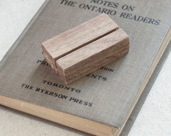 May include: A small, rectangular wooden book holder with a slot cut into the top. The holder is made of light brown wood and sits on top of a vintage book with gray cover and black text. The book's title is "Notes on the Ontario Readers."