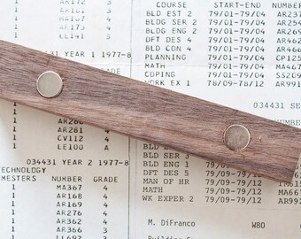May include: A wooden ruler with two silver magnets attached to it. The ruler is lying on top of a sheet of paper with text and numbers. The text on the paper includes course names, dates, and numbers. The ruler is approximately 30 cm long.