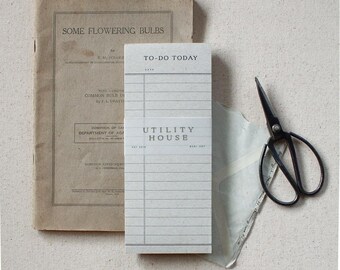 May include: A vintage book titled "Some Flowering Bulbs" and a notepad with the text "To-Do Today" and "Utility House" are arranged with black scissors and a translucent paper envelope. The items are set against a neutral background, creating a classic aesthetic.