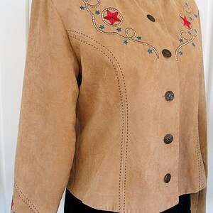 May include: Tan suede jacket with embroidered red stars and blue star accents. The jacket features a stand-up collar, decorative stitching, and bronze-colored buttons. A western-themed garment.