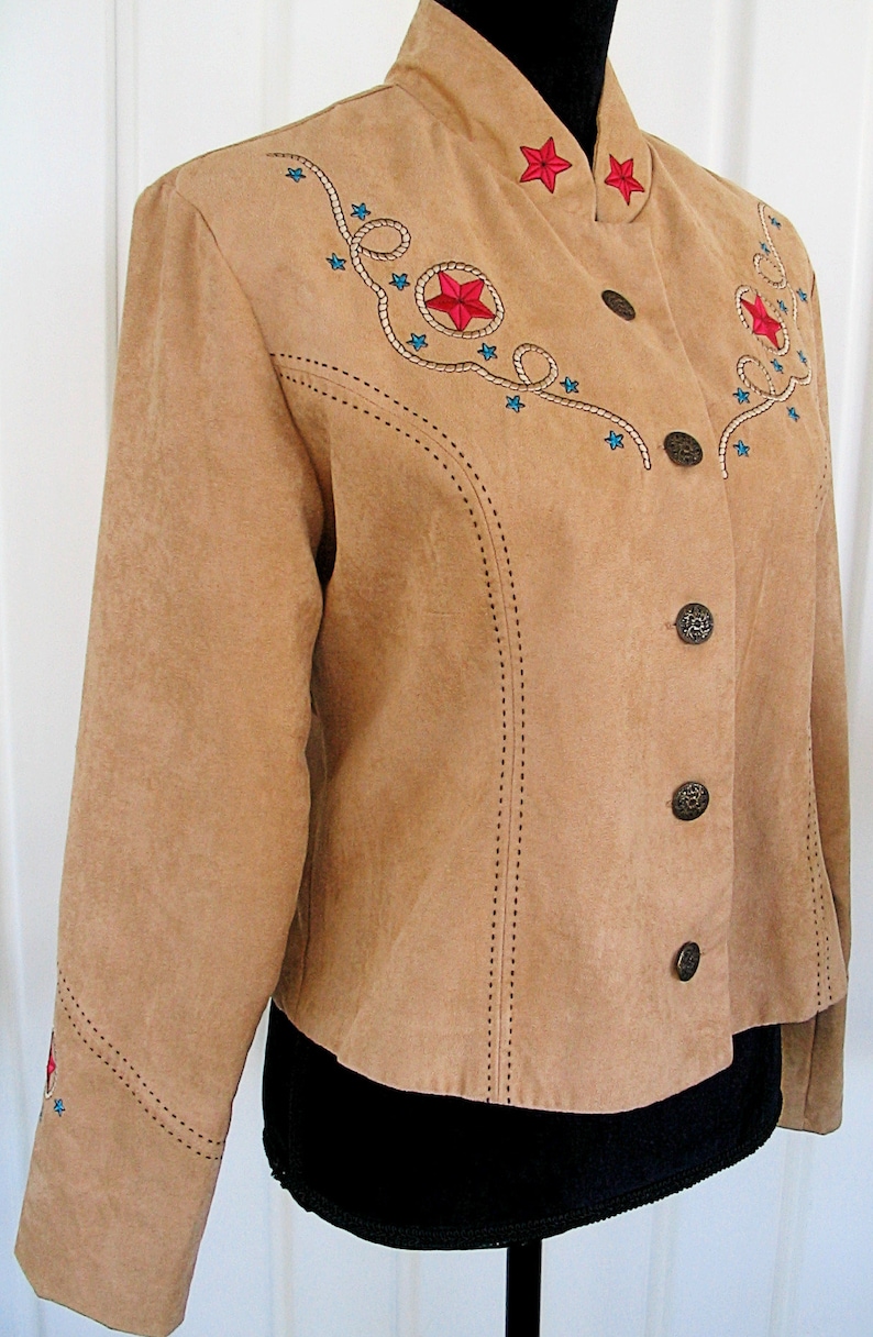 May include: Tan suede jacket with embroidered red stars and blue star accents. The jacket features a stand-up collar, decorative stitching, and bronze-colored buttons. A western-themed garment.