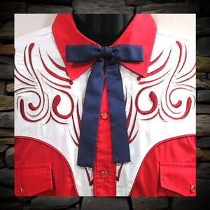 May include: A white shirt with red accents, a red collar, and two red pockets. The shirt features a navy blue bow tie and decorative stitching. The shirt has red buttons and is displayed on a black mannequin.