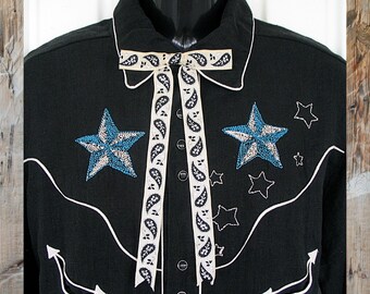 May include: A black long-sleeve shirt featuring white piping, two large blue and silver star appliqués, and smaller embroidered star outlines. A cream-colored ribbon with a black paisley pattern is tied at the collar, and white arrow-shaped accents.