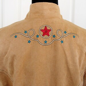 May include: Tan suede-like jacket with embroidered design on the back. The design features a red star within a rope circle, blue stars, and a rope-like pattern. The jacket has a high collar and stitched details.