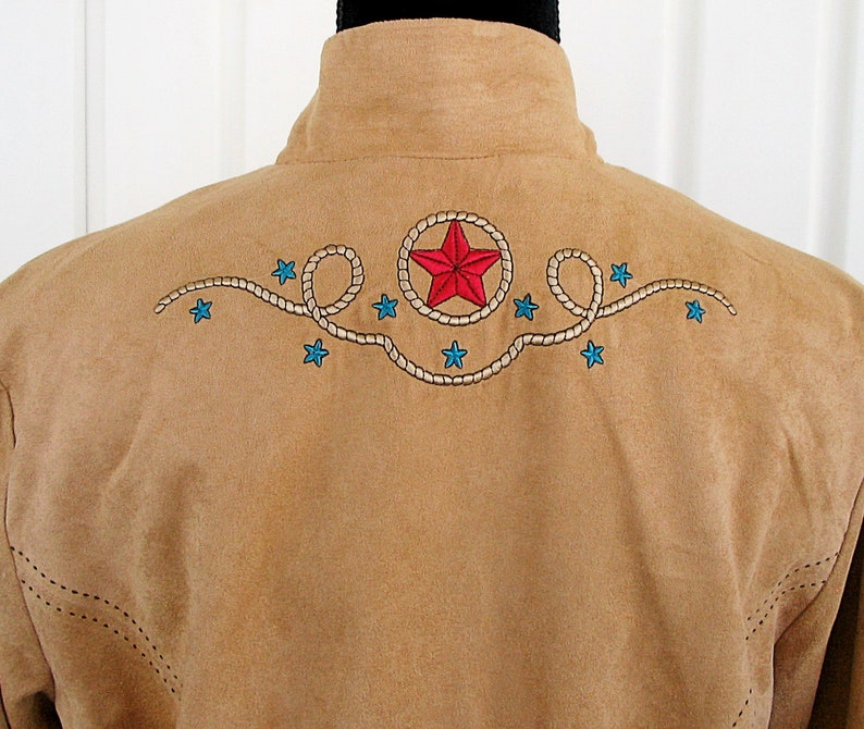 May include: Tan suede-like jacket with embroidered design on the back. The design features a red star within a rope circle, blue stars, and a rope-like pattern. The jacket has a high collar and stitched details.