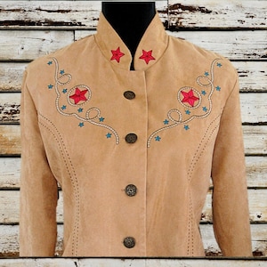 May include: Tan suede jacket with red star and blue starburst embroidery. The jacket has a high collar, decorative buttons, and a Western-inspired design. The jacket is displayed on a black mannequin.