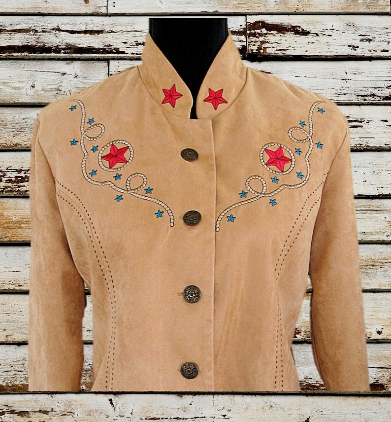 May include: Tan suede jacket with red star and blue starburst embroidery. The jacket has a high collar, decorative buttons, and a Western-inspired design. The jacket is displayed on a black mannequin.