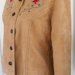 May include: Tan suede jacket with a stand-up collar and button closure. The jacket features embroidered red stars, blue stars, and rope detailing. The sleeves and collar are also decorated with star designs. The jacket is displayed on a black mannequin.