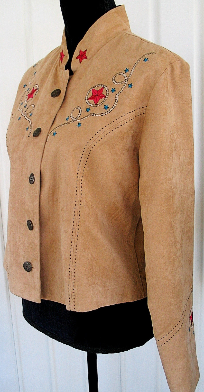 May include: Tan suede jacket with a stand-up collar and button closure. The jacket features embroidered red stars, blue stars, and rope detailing. The sleeves and collar are also decorated with star designs. The jacket is displayed on a black mannequin.