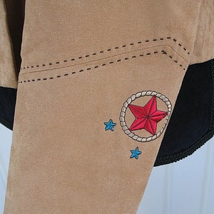 May include: Tan suede jacket detail with embroidered red star inside a rope circle, and two small blue stars. The jacket has black trim and decorative stitching.