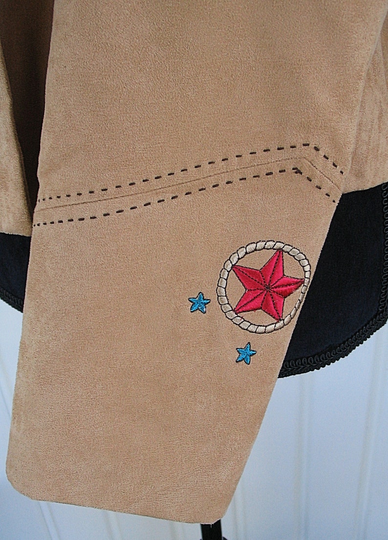 May include: Tan suede jacket detail with embroidered red star inside a rope circle, and two small blue stars. The jacket has black trim and decorative stitching.