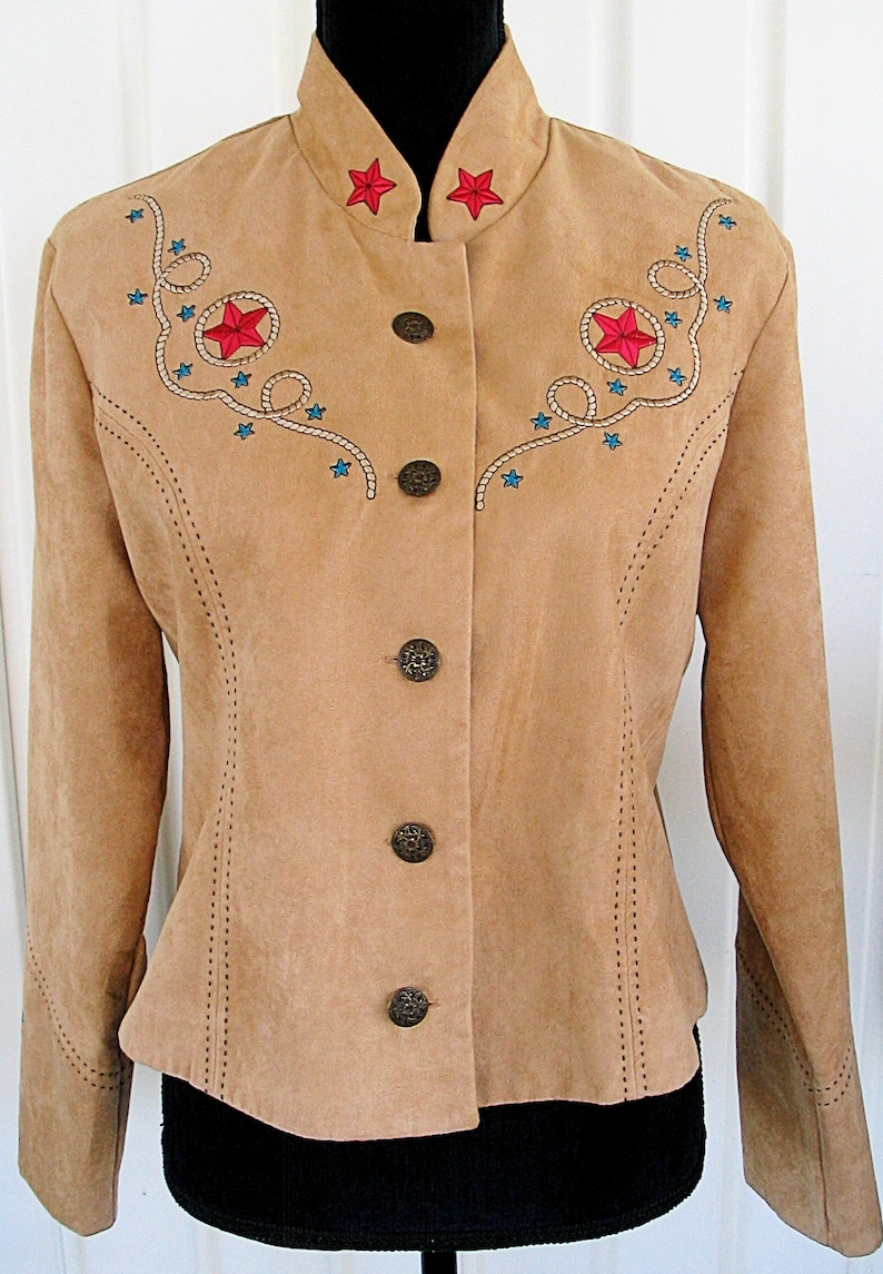 May include: Tan suede jacket with a mandarin collar and decorative stitching. Features embroidered red stars and turquoise accents with rope detailing. The jacket has four decorative buttons.