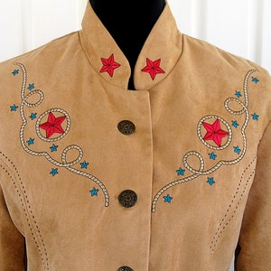May include: A tan suede jacket with a mandarin collar and three decorative buttons. The jacket features embroidered red stars, blue stars, and rope detailing. The jacket has a western theme.