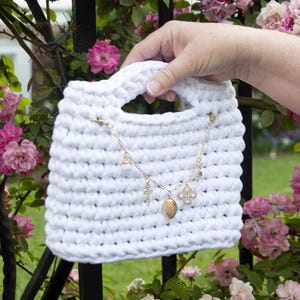 May include: A white crochet tote bag with a gold chain and charms. The bag has a rectangular shape with a handle. The gold chain features a cross, a locket, and a four-leaf clover charm. The background includes pink roses.